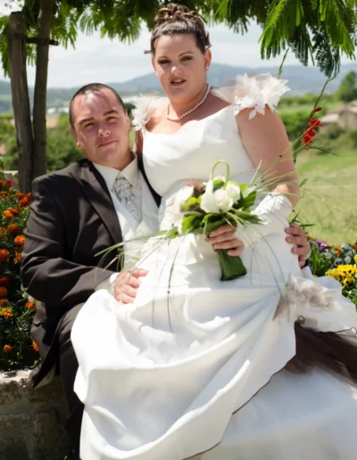 Un couple de mariés pose à lextérieur par une journée ensoleillée. La mariée tient un bouquet de verdure, porte une robe blanche avec un corsage en dentelle, tandis que le marié porte un costume sombre. Ils sont entourés de fleurs colorées et dune végétation luxuriante.