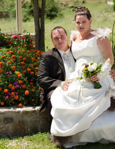 Un couple de mariés pose pour une photo dans un jardin en plein air. La mariée, en robe blanche, tient un bouquet tout en étant assise à côté du marié, qui porte un costume sombre. Ils sont entourés de fleurs et de verdure aux couleurs vives.