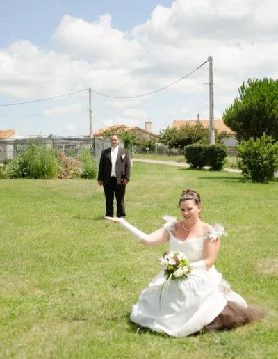 Une mariée en robe de mariée est à genoux sur lherbe, tenant un bouquet, et fait des gestes ludiques de la main. À larrière-plan, un homme en costume se tient dans un champ vert ouvert sous un ciel partiellement nuageux.