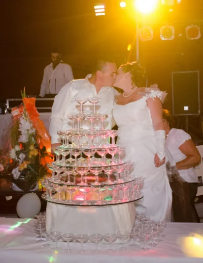Un couple de mariés sembrasse à côté dune pyramide de flûtes de champagne vides, sous un éclairage doux et coloré. La scène comprend des compositions florales et un DJ en arrière-plan.