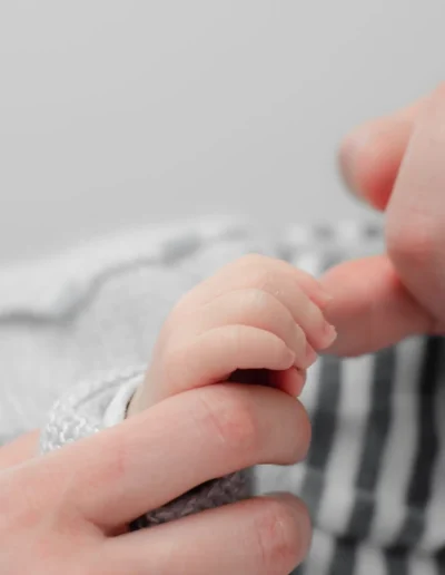 Gros plan dune main dadulte tenant doucement la main dun bébé. Le bébé porte une tenue rayée grise et blanche, et ladulte touche les petits doigts du bébé sur un fond doux et flou.