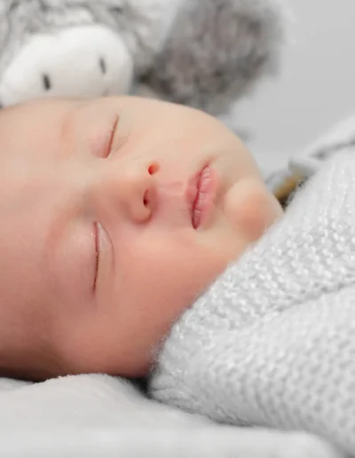 Un bébé endormi paisiblement dans une tenue en tricot gris doux, allongé sur une couverture blanche. Un animal en peluche gris est blotti à proximité, ajoutant une touche douillette et sereine à limage.