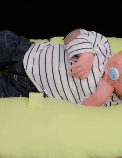 Un bébé est allongé sur un coussin vert clair, vêtu dune chemise rayée, dun jean et de baskets grises. Le bébé tient une tétine bleue dans sa bouche et a une expression douce sur un fond noir.
