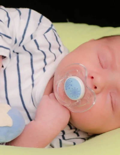 Un bébé endormi, vêtu dune tenue rayée, tient une tétine bleue dans sa bouche. Le bébé est allongé sur un coussin vert clair et a une peluche bleue toute douce à côté de lui.