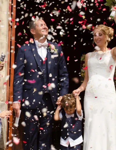 Un couple de jeunes mariés se tient joyeusement sous une pluie de pétales de fleurs le jour de leur mariage. La mariée lève son bouquet en signe de célébration. Deux enfants se tiennent à leurs côtés, lun deux se couvrant les oreilles. Des invités aux tenues colorées les entourent, immortalisant linstant.