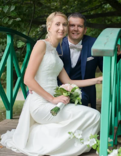 Une mariée en robe blanche est assise sur un pont vert, tenant un bouquet, à côté dun marié en costume bleu avec un nœud papillon. Ils sourient, entourés dune végétation luxuriante.