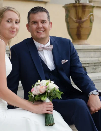 Un couple de mariés sont assis ensemble sur des marches en pierre. La mariée porte un bouquet de fleurs blanches et roses et porte une robe en dentelle blanche. Le marié porte un costume bleu foncé avec un nœud papillon. Ils sourient tous les deux à la caméra.
