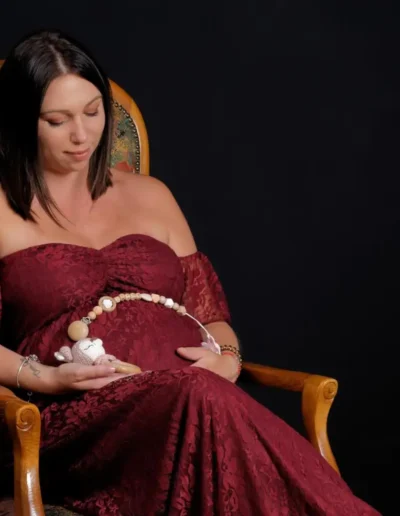 Une femme vêtue dune robe en dentelle bordeaux est assise sur une chaise en bois sur un fond sombre. Elle tient une petite peluche et un collier de perles décoratives, quelle pose délicatement sur son ventre de femme enceinte. Son expression est calme et sereine.