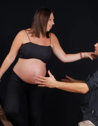 Une femme enceinte en tenue noire sappuie sur un escabeau, tenant la main dun homme aux cheveux courts qui se dirige vers elle. Le fond est noir.