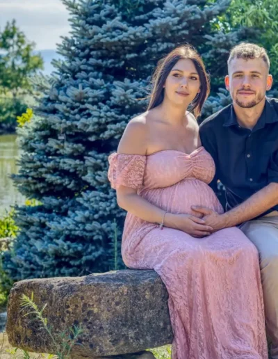 Une femme enceinte en robe rose est assise sur un banc de pierre à côté dun homme en chemise noire et pantalon beige. Ils sont dehors près dun lac, entourés de verdure luxuriante et darbres, sous un ciel bleu clair.