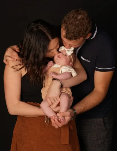 Un couple enlace avec amour son nouveau-né, qui porte un bandeau à nœud, sur un fond sombre. La femme embrasse le bébé sur la joue tandis que lhomme embrasse la tête du bébé, créant ainsi un moment de tendresse en famille.