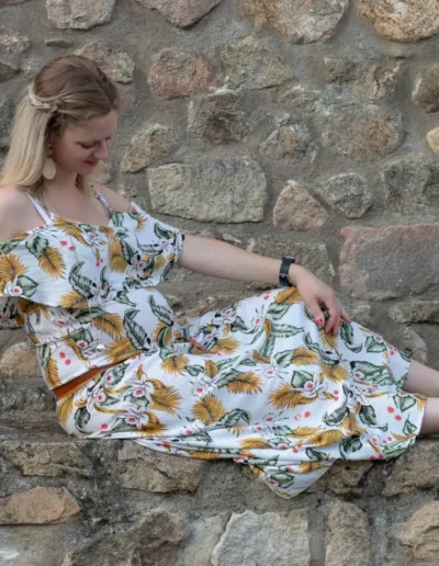 Une femme en robe fleurie est assise sur un mur de pierre, regardant vers le bas et souriant. Larrière-plan est une surface en pierre texturée, créant une atmosphère rustique. Elle porte des sandales et un bracelet.