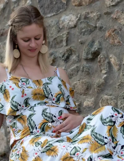 Une femme enceinte vêtue dune robe fleurie est assise contre un mur de pierre, les yeux baissés et touchant doucement son ventre. Elle a les cheveux longs jusquaux épaules et porte de grandes boucles doreilles rondes.