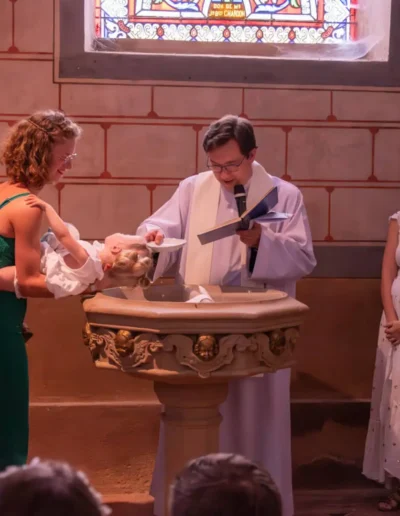 Cérémonie de baptême dans une église. Un prêtre en robe blanche tient un livre, tandis quun couple soutient un bébé au-dessus dun bénitier. Deux parrains et marraines se tiennent à proximité. Un vitrail est visible à larrière-plan.