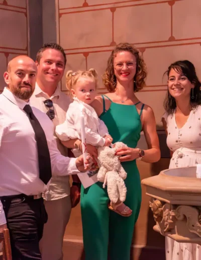 Un groupe de cinq adultes et un enfant se tiennent devant des fonts baptismaux en pierre dans une pièce joliment décorée. Les adultes sont habillés en tenue de soirée et lenfant est tenu dans les bras de lun deux, vêtu de vêtements de couleur claire.