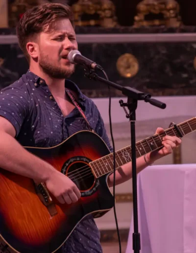 Un homme chante avec passion dans un micro tout en jouant de la guitare acoustique sur scène. Il a les cheveux courts et porte une chemise boutonnée à manches courtes de couleur foncée. Larrière-plan présente des éléments décoratifs et une nappe dautel blanche.