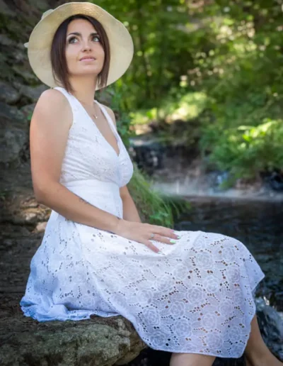 Une femme vêtue dune robe en dentelle blanche et dun chapeau de paille est assise près dun petit ruisseau dans un décor de forêt verdoyante et luxuriante, le regard pensif vers le haut. La lumière du soleil filtre à travers les arbres, créant une atmosphère sereine et paisible.