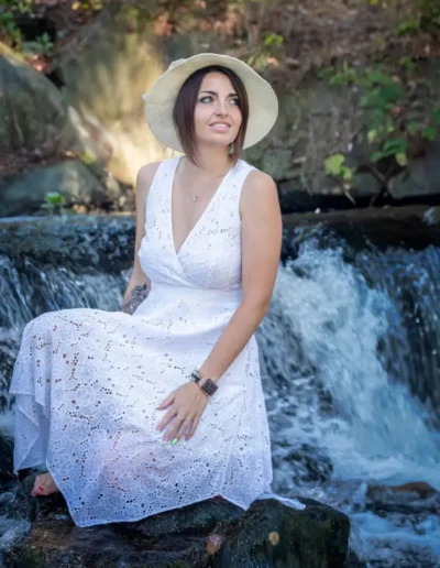 Une femme vêtue dune robe en dentelle blanche et dun chapeau de paille est assise sur un rocher près dune petite cascade. Elle regarde vers le côté, entourée deau courante et de verdure.