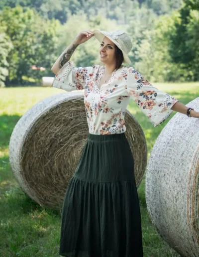 Une femme portant un chemisier fleuri et une jupe longue pose avec sa main sur une botte de paille dans un champ ensoleillé, portant un chapeau et souriant. Des arbres verts luxuriants et dautres bottes de paille sont à larrière-plan.