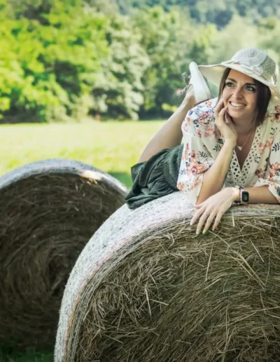 Une femme portant un chemisier fleuri et un chapeau est allongée sur une grosse botte de foin dans un champ ensoleillé. Elle sourit et pose son menton sur sa main. Larrière-plan est luxuriant avec des arbres verts et de lherbe, créant une atmosphère sereine et rurale.