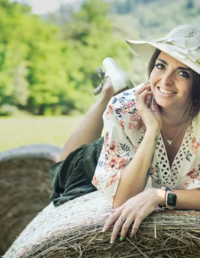 Une femme portant un chemisier fleuri, un short et un chapeau de soleil est allongée sur une botte de foin ronde dans un champ herbeux avec des arbres en arrière-plan. Elle sourit et pose son menton sur sa main.