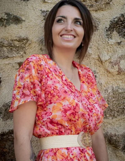 Une femme aux cheveux bruns courts sourit debout contre un mur de pierre. Elle porte une robe fleurie colorée avec une large ceinture beige ornée dune boucle décorative.