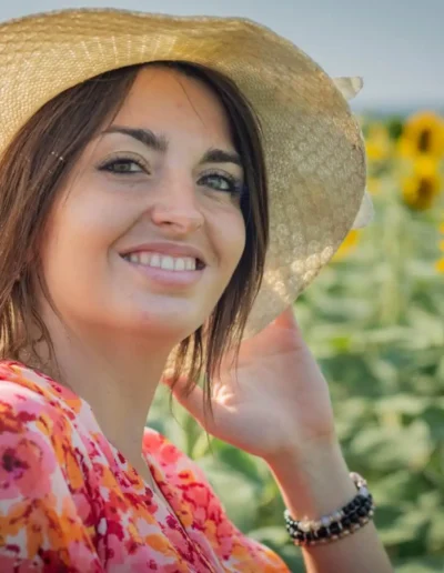 Une femme portant un chapeau de paille et une robe fleurie sourit dans un champ de tournesols. Les tournesols fleurissent autour delle sous un ciel bleu clair.