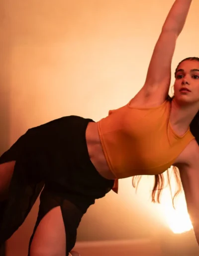 Une danseuse vêtue dun haut court orange et dune jupe noire exécute une pose de planche latérale sur scène. Léclairage chaleureux crée une ambiance dramatique alors quelle regarde vers le haut et quune autre danseuse est partiellement visible en arrière-plan.