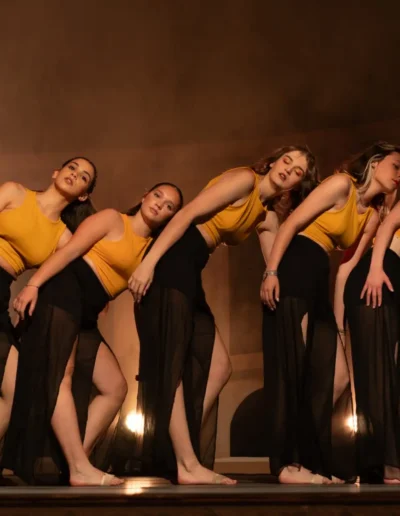 Cinq danseurs en haut jaune et jupe noire se produisent dans un décor faiblement éclairé. Ils se tiennent en rang, appuyés les uns contre les autres dans une pose expressive. Des projecteurs sont visibles en arrière-plan, projetant une lumière chaleureuse.