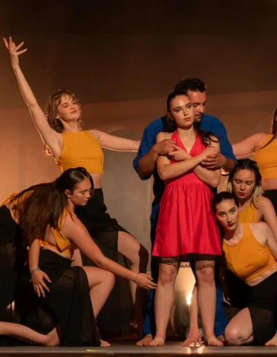 Un groupe de six danseurs se produit sur scène. Un danseur central en robe rouge est entouré dautres danseurs en haut jaune et jupe noire. Un homme en bleu tient le personnage central. Léclairage est chaleureux, mettant en valeur leurs poses expressives.