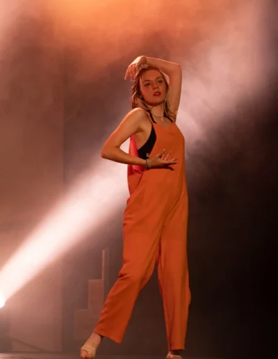Une personne en combinaison orange danse sur scène, prenant une pose équilibrée avec un bras au-dessus de la tête. Elle est illuminée par un éclairage dramatique, avec des effets de fumée créant une atmosphère dynamique.