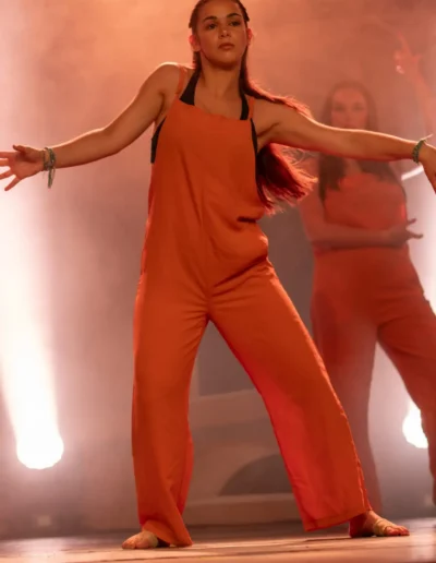 Une personne en combinaison orange exécute une danse sur scène, les bras tendus. Elle est pieds nus, avec un autre danseur en arrière-plan. Un éclairage doux et chaleureux et de la brume créent une atmosphère dramatique.