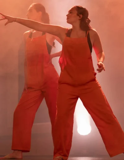 Deux danseurs en combinaison orange assortie se produisent sur scène devant un arrière-plan enfumé et rétroéclairé. Lun des danseurs pointe vers lavant de manière spectaculaire tandis que lautre se tient derrière, tous deux immergés dans une lumière chaleureuse.