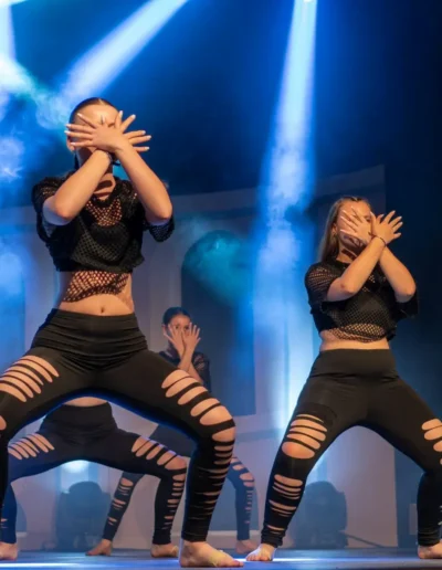Un groupe de danseurs se produit avec énergie sur scène sous des lumières vives. Ils portent des tenues noires avec des hauts en maille et des leggings déchirés, prenant une pose dramatique avec les mains couvrant leur visage. Des effets de fumée renforcent latmosphère vibrante.