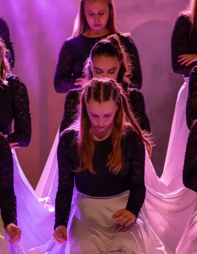 Un groupe de filles en hauts noirs à manches longues et jupes blanches flottantes se produit sur scène sous un éclairage violet. La fille la plus proche est à genoux, la tête légèrement penchée, tandis que dautres se tiennent derrière elle dans une pose chorégraphiée.