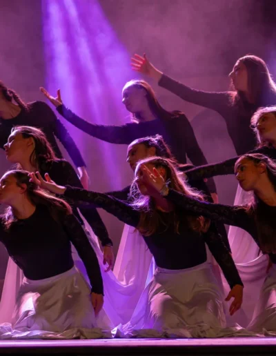 Un groupe de danseuses en jupe longue se produit sur scène sous un éclairage violet, adoptant des poses expressives avec les bras tendus. Le fond est brumeux, renforçant latmosphère dramatique.