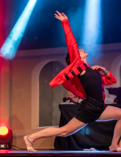Une danseuse en veste rouge prend une pose dramatique sur scène sous des lumières colorées. Deux autres interprètes en tenue rouge sont à larrière-plan, lun debout et lautre accroupi. Larrière-plan présente un motif darche.