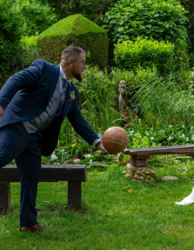 Un marié joue avec un ballon de basket sur son pied, debout sur un banc dans un jardin. La mariée, vêtue dune robe blanche, réagit avec surprise, tenant un bouquet. La verdure luxuriante et un étang forment la toile de fond.