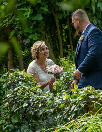 Une mariée en robe blanche tenant un bouquet sourit à un marié en costume bleu marine. Ils sont entourés dun feuillage vert luxuriant et de fleurs dans un décor de jardin.