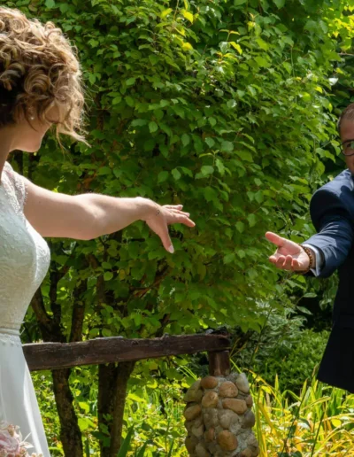 Un couple de mariés se tendent la main sur un pont en bois entouré dune végétation luxuriante. La mariée tient un bouquet et porte une robe en dentelle, tandis que le marié porte un costume bleu avec des lunettes.