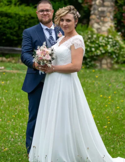 Une mariée en robe blanche tient un bouquet et se tient debout à côté dun marié en costume bleu avec une cravate. Ils sont à lextérieur sur une zone herbeuse avec des arbres et une structure en pierre en arrière-plan.
