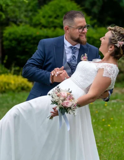 Un marié soulève joyeusement la mariée dans un jardin. La mariée tient un bouquet de fleurs roses et blanches et sourit, tout en portant une robe blanche à manches en dentelle. Le marié porte un costume bleu marine avec une cravate à motifs, tous deux partageant un moment de joie.