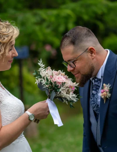 Un couple se tient dans un jardin verdoyant. La femme, vêtue dune robe en dentelle blanche, tient un bouquet de fleurs roses et blanches. Lhomme, vêtu dun costume bleu, se penche en avant, sourit et sent les fleurs.