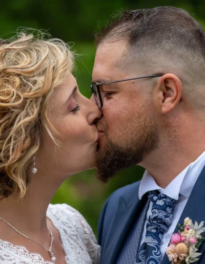 Un couple sembrasse en plein air. La femme a les cheveux blonds bouclés et porte une robe en dentelle, tandis que lhomme a les cheveux courts, des lunettes et est vêtu dun costume avec une cravate et une boutonnière. La verdure est floue à larrière-plan.
