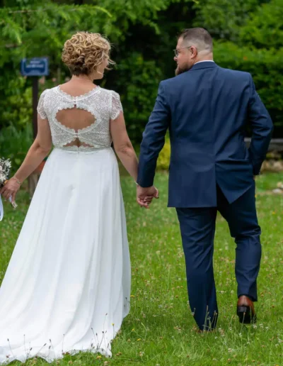 Un couple marche main dans la main sur un chemin herbeux. La femme porte une robe de mariée blanche et lhomme est en costume bleu marine. Ils sont photographiés de dos, entourés de verdure.
