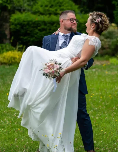 Dans un jardin fleuri et verdoyant, un marié en costume bleu soulève avec enjouement la mariée, qui tient un bouquet et porte une robe de mariée blanche à manches en dentelle. Ils se sourient tous les deux avec amour.