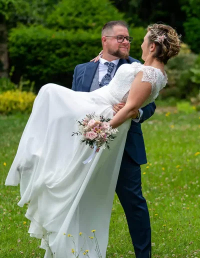 Un marié en costume bleu porte joyeusement sa mariée dans une robe de mariée en dentelle blanche sur un champ herbeux. Elle tient un bouquet de fleurs roses et blanches. Le couple semble heureux, avec de la verdure en arrière-plan.