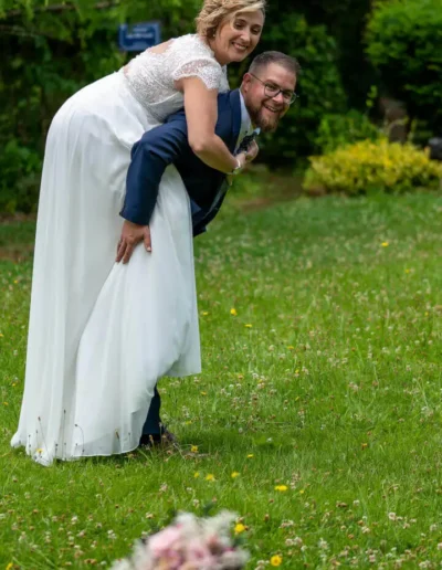 Dans un jardin, une mariée chevauche joyeusement le dos du marié. Ils sourient tous les deux, la mariée porte une robe blanche et le marié un costume bleu marine. Un bouquet est flou au premier plan et la verdure les entoure.