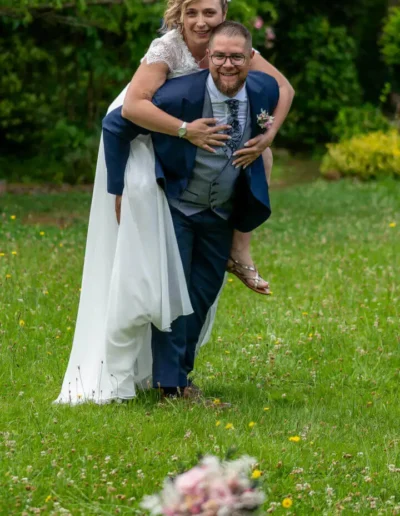 Un couple de mariés se trouve dans un jardin, la mariée chevauchant joyeusement le dos du marié. Tous deux sourient et sont habillés en tenue de mariage. Un bouquet est visible sur lherbe au premier plan.