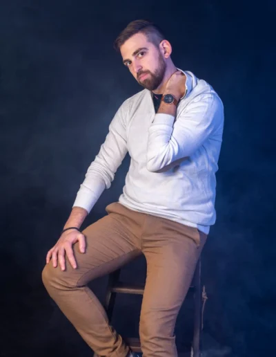 Un homme avec une barbe et des cheveux courts est assis sur un tabouret dans une pose décontractée sur un fond sombre et enfumé. Il porte un pull gris clair, un pantalon marron et des baskets blanches. Sa main droite repose sur son genou, tandis que sa main gauche touche son cou.